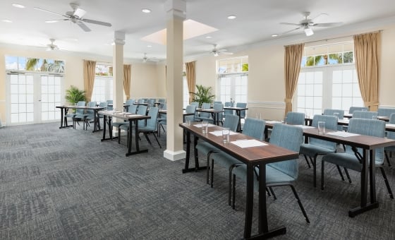 Conference room with long wooden tables arranged in rows set with paper, pen and empty water glass. Blue chairs sit at each spot.